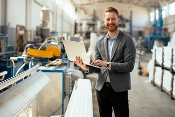Manufacturing CFOs A man in business clothes inside of a manufacturing facility holding a laptop and smiling.