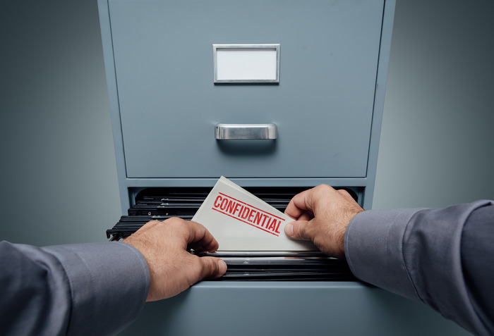 Hands slipping a folder marked Confidential into a filing cabinet drawer.