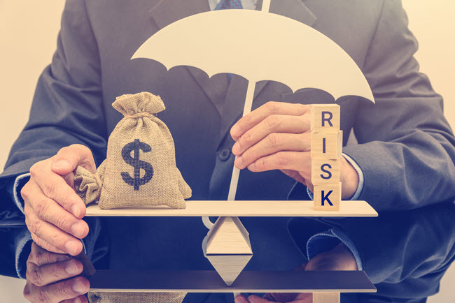 Hands holding an umbrella over a money bag balanced against blocks spelling Risk.