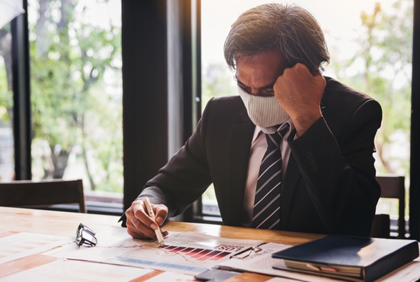 A masked businessman on the phone while reviewing paperwork at his desk.
