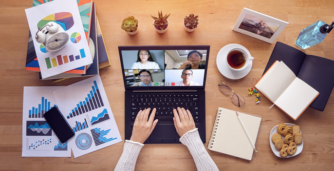 Overhead view of a remote worker on a video call at a laptop surrounded by charts and coffee.