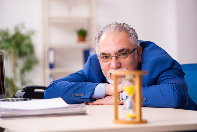 An older man resting his head on his desk beside an hourglass.