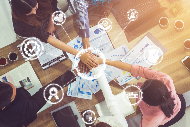 Overhead view of a team joining hands over a desk covered in documents and product icons.