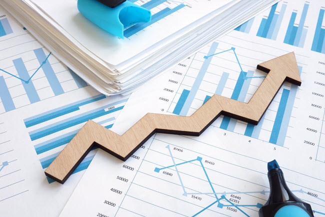 A wooden arrow trending upward resting on printed bar charts and financial reports.