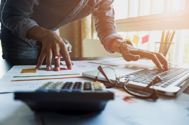 A person working at a laptop with printed reports, a calculator and charts on the desk.