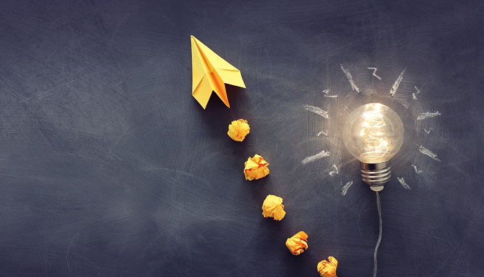 A trail of crumpled paper balls leading to a single glowing lightbulb.