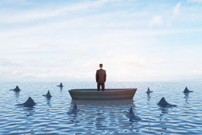 A lone businessman standing in a small boat surrounded by circling shark fins.