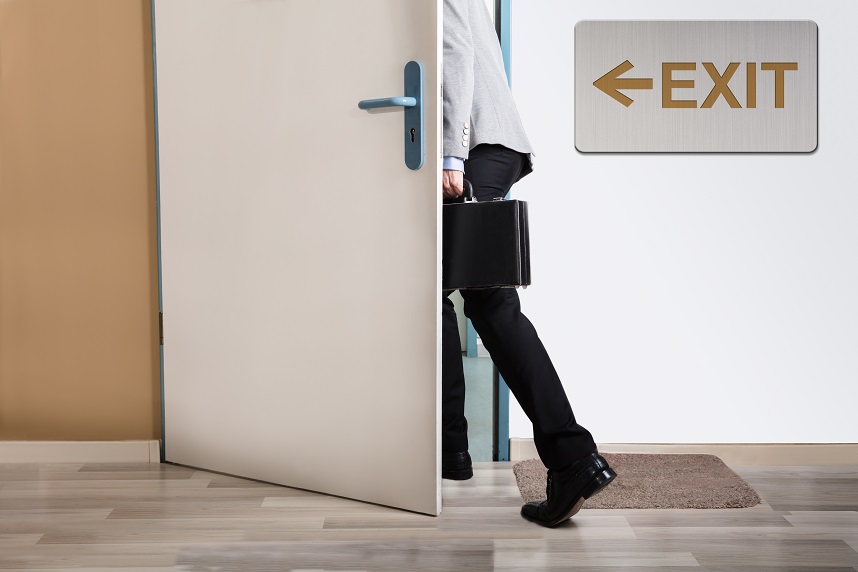 A person carrying a briefcase walking out a door beneath an exit sign.