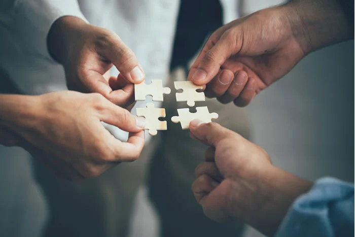Four sets of hands fitting puzzle pieces together to complete a puzzle.