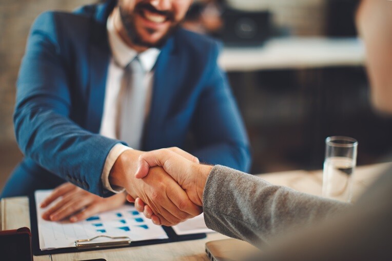 Two people shaking hands across a table of financial documents after closing a deal.