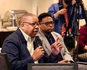 Raphael Irving speaking into a microphone at a podium alongside another presenter.