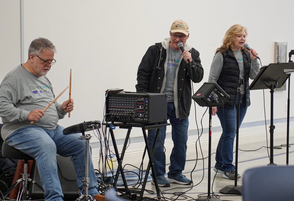 Three volunteers in Community Meals shirts perform live music, one on drums and two singing into microphones.