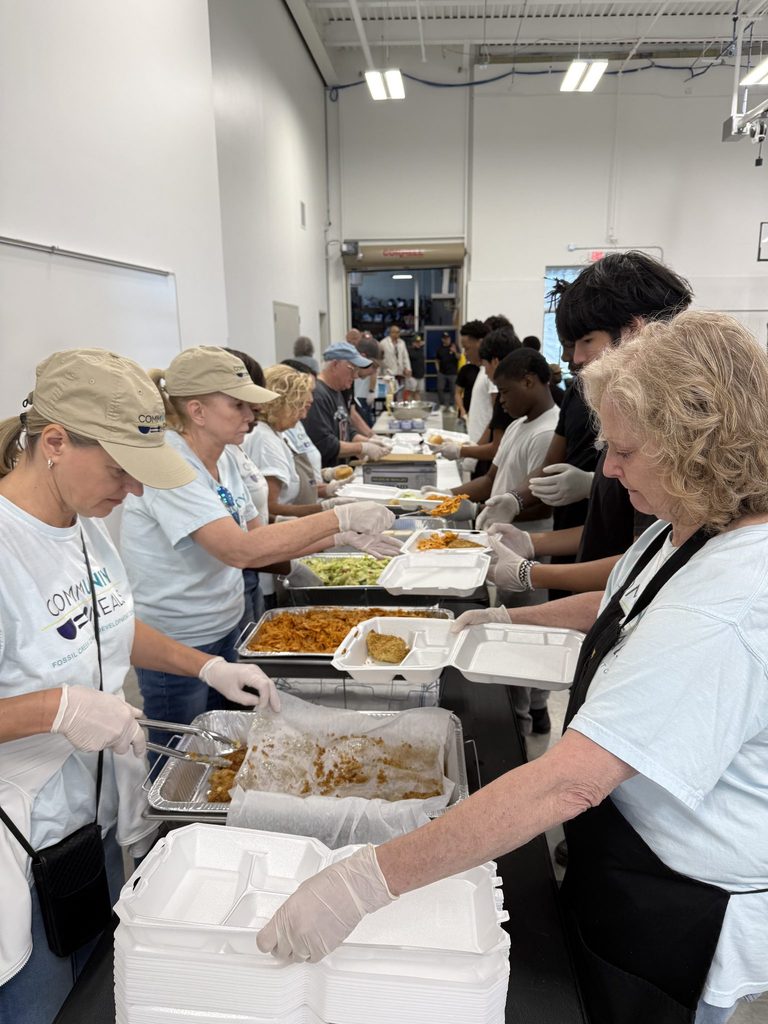 Community Meals volunteers in gloves and caps fill takeout containers along a serving line in a warehouse kitchen.