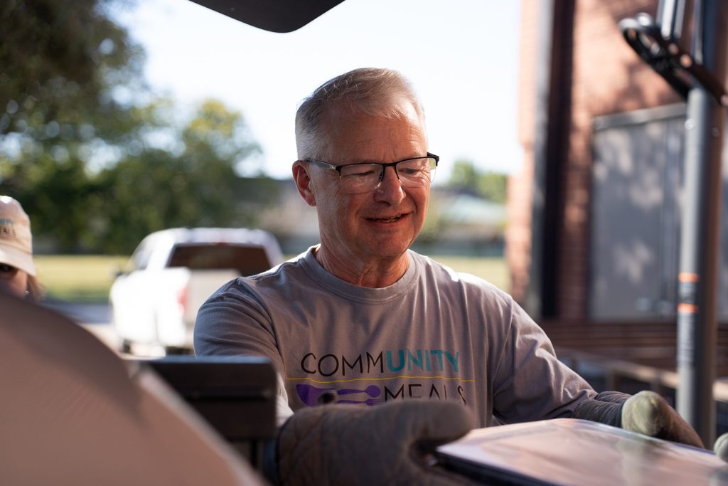 Randy Teuber smiles as he works an outdoor grill in oven mitts, wearing a Community Meals volunteer shirt.