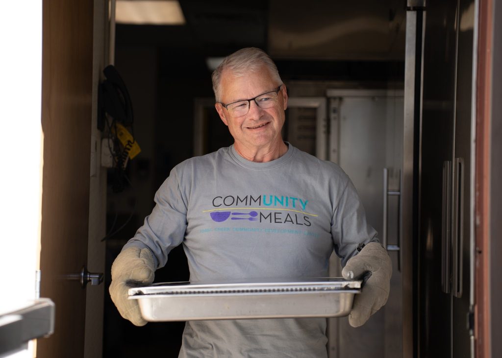 Randy Teuber carries a sheet pan through a commercial kitchen doorway in a Community Meals volunteer shirt.