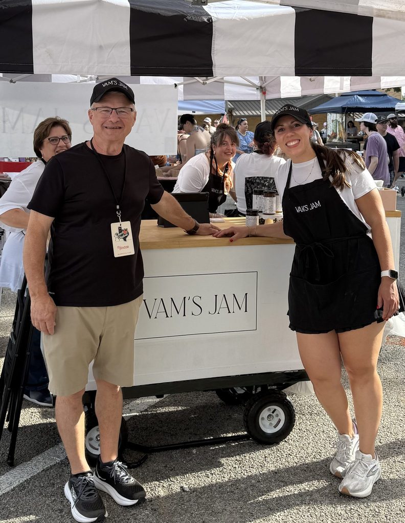 Randy Teuber stands with a vendor beside the Vam's Jam cart at an outdoor farmers market under a striped canopy.
