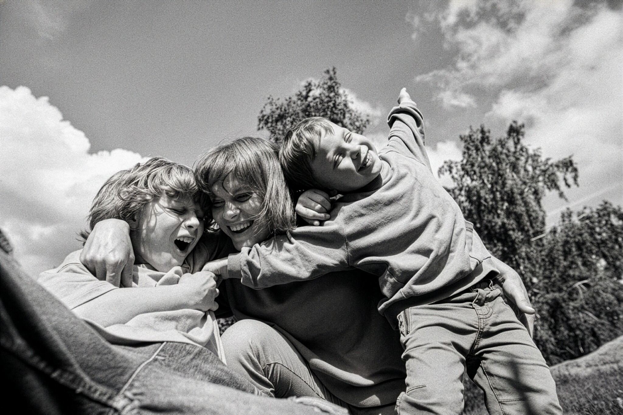 Laughing woman hugs two boys as they pile onto her outdoors under a sunny sky