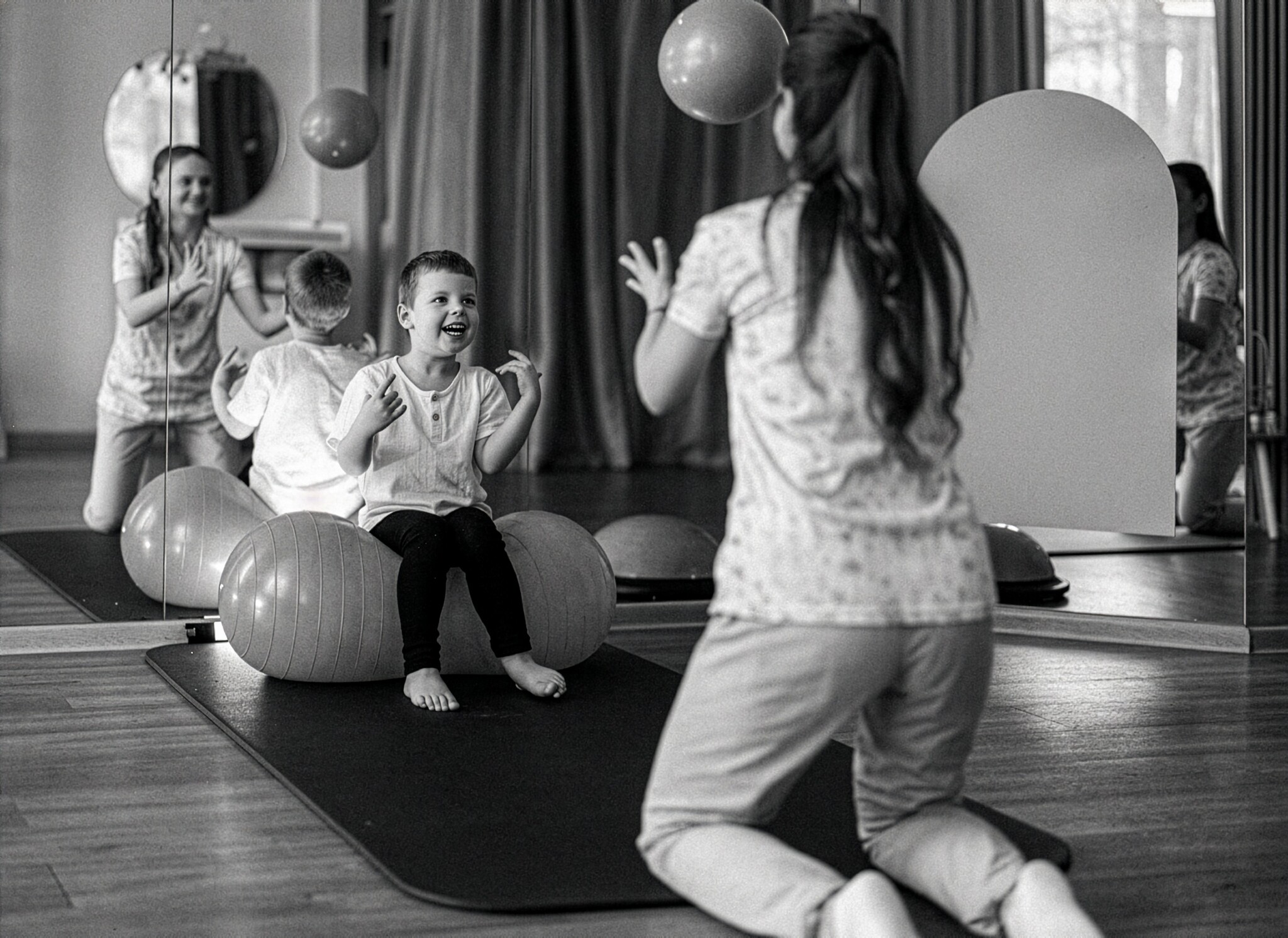 Smiling boy sits on a peanut ball and reaches to catch a ball tossed by a therapist in a mirrored therapy room